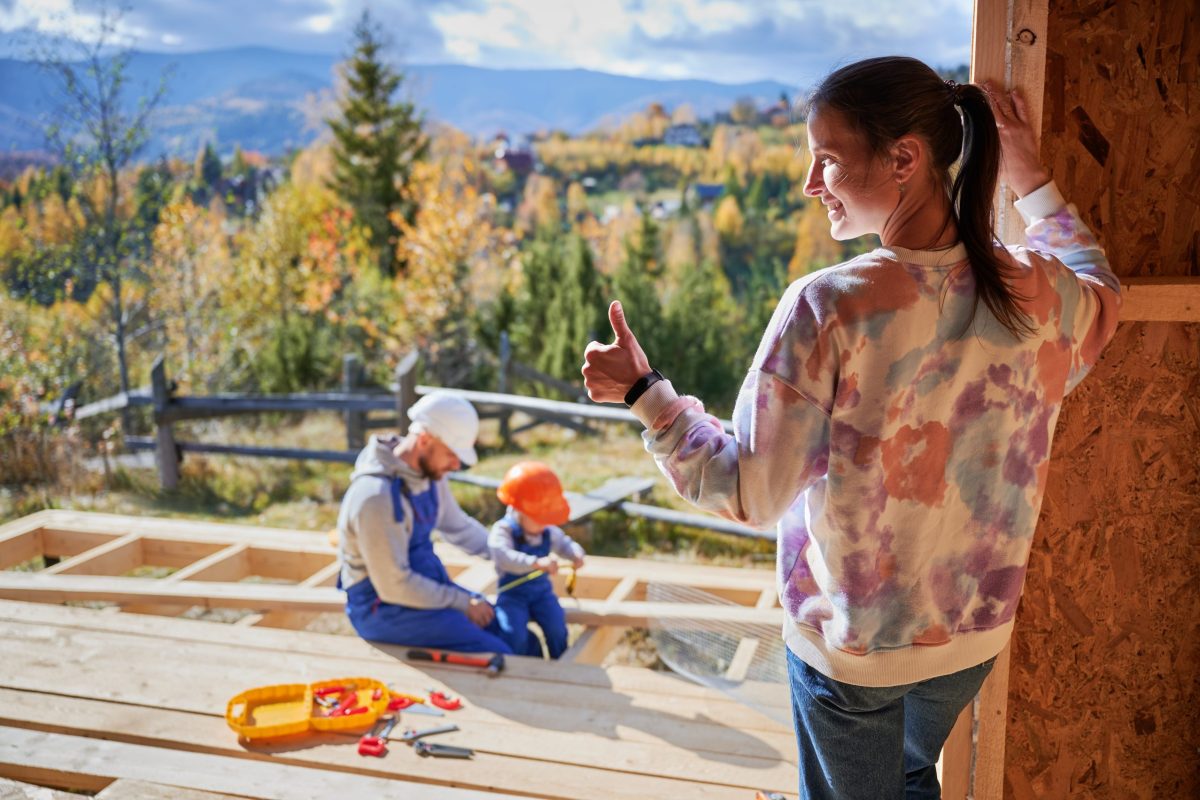 Father, mother and son building wooden frame house. Toddler boy helping his daddy, while woman looking for them on construction site. Guys wearing helmet and blue overalls. Carpentry, family concept.